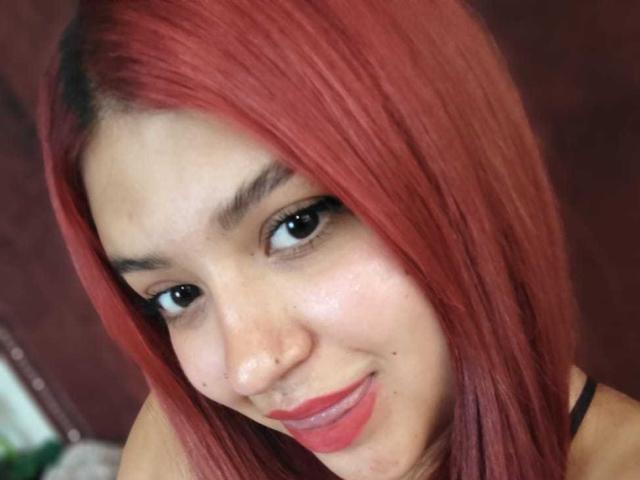 Close-up photo of AmberCutel with vibrant red hair, black eyeliner, and red lipstick. She's looking directly at the camera with a slight smile, showing a small facial mole. Fucking hot.