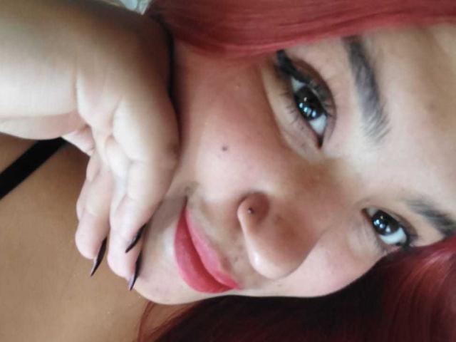 Close-up of AmberCutel with red hair, fair skin, and black eyeliner, looking directly at the camera with a slight smile. Hand with long nails touches her chin. Fucking sexy.