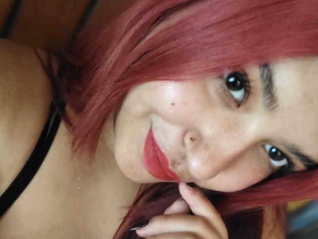 Close-up photo of AmberCutel with red hair, black eyeliner, and red lipstick, looking seductively at the camera with a finger near her lips. Her eyes are slightly glazed. She's wearing a black strap.