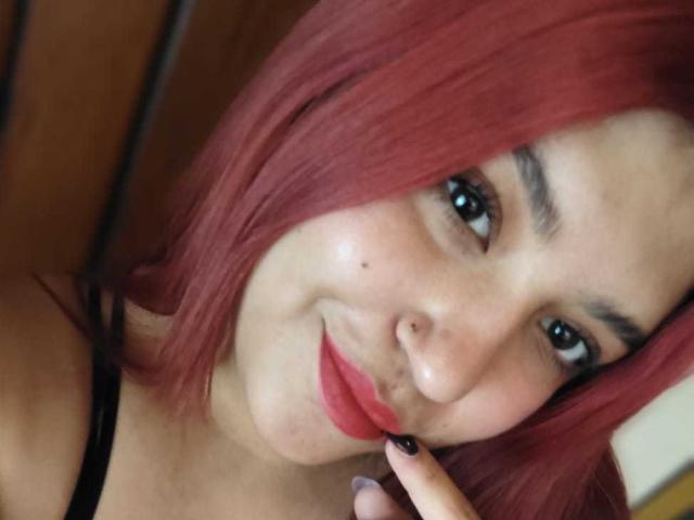 Close-up photo of AmberCutel with red hair, black eyeliner, and red lipstick, touching her lip with a finger, brown eyes, light skin, subtle freckles, wearing a black strap. Fucking sexy.