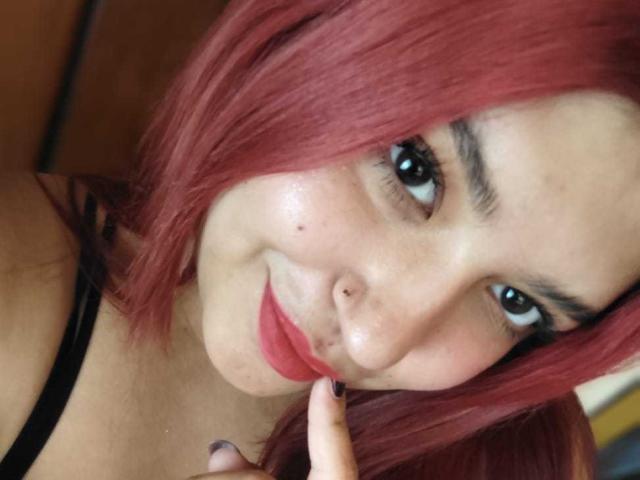 Autofoto de primer plano de AmberCutel con cabello rojo, delineador de ojos negro y lápiz labial rojo, sonriendo mientras toca sus labios. dedo. Sus grandes ojos brillan ligeramente. Su piel es clara..