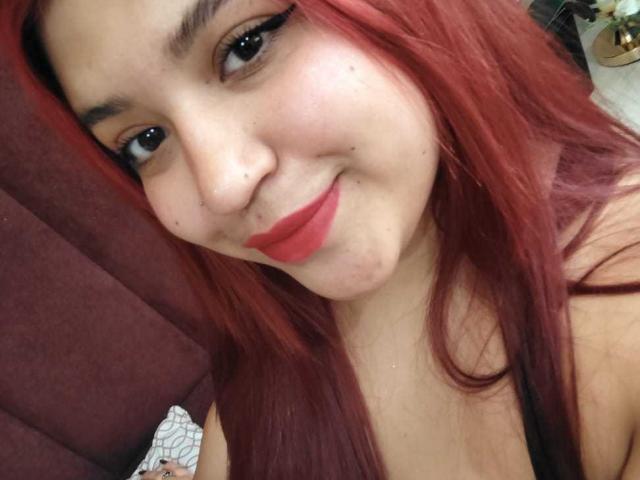 Close-up selfie van AmberCutel met lang rood haar, dikke zwarte eyeliner en felrode lipstick. Ze glimlacht lichtjes en draagt een klein neuspiercing. Haar decolleté is zichtbaar, wat suggereert dat ze een vol figuur heeft..