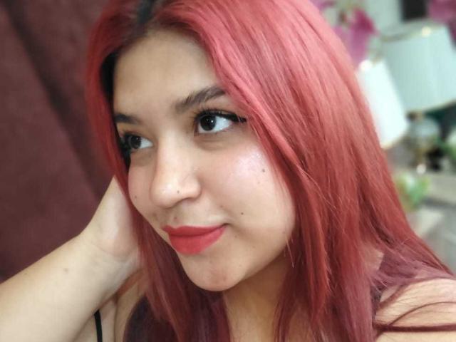 Nahaufnahme von AmberCutel mit hellroten Haaren, schwarzem Eyeliner und rotem Lippenstift, die zur Seite schaut und mit der Hand ihr Gesicht berührt. Gesicht. Unschärfer Hintergrund mit weicher Beleuchtung. Leices Lächeln, verführerischer Ausdruck..