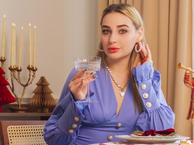 Barbara Rosy, avec de longs cheveux blonds, une robe bleue, tenant un verre de vin, regardant la caméra, séduisante, élégante, rouge. rouge à lèvres, collier de perles, chandeliers et sapin de Noël en arrière-plan.