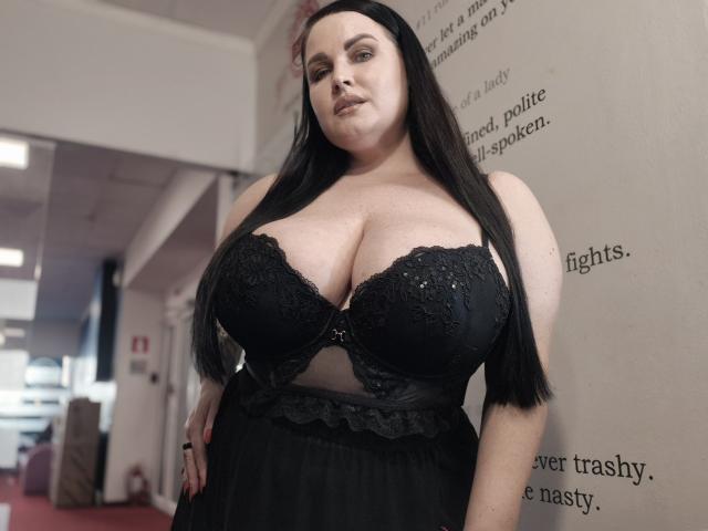 Photograph of a plus-sized woman with long black hair, wearing a black lace bra that accentuates her large breasts. She stands against a wall with text, looking seductively at the camera. Her outfit is tight and revealing.