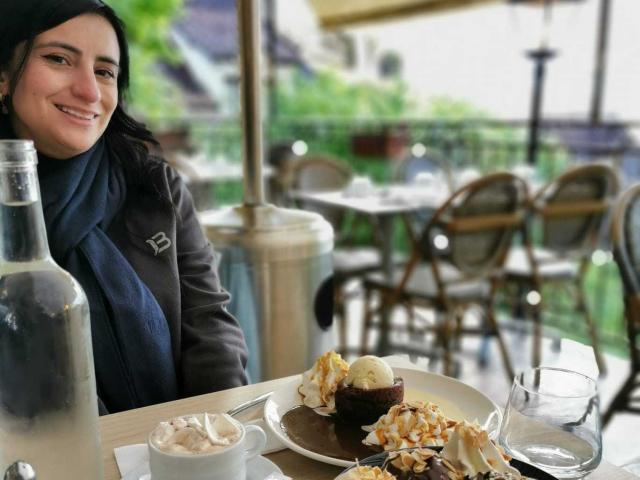 Brenda Cantorel, aux cheveux foncés, souriante, portant un manteau noir, est assise à une table d'un café en plein air, avec une assiette de dessert. crème et des noix. Des bouteilles et des chaises floues en arrière-plan. Tellement mignon..