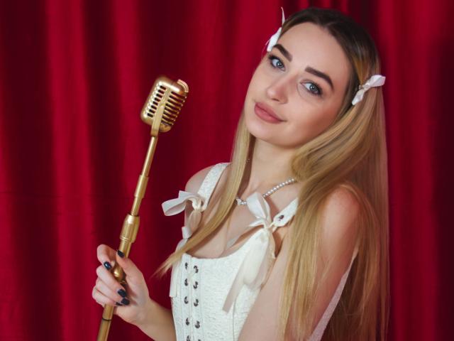 BarbaraRosy, una donna bionda con occhi azzurri, che tiene un microfono dorato, indossa un corsetto bianco con dettagli a fiocco, rosso sfondo con tenda, capelli lunghi e lisci, mollette bianche, leggero sorriso, posa sensuale.