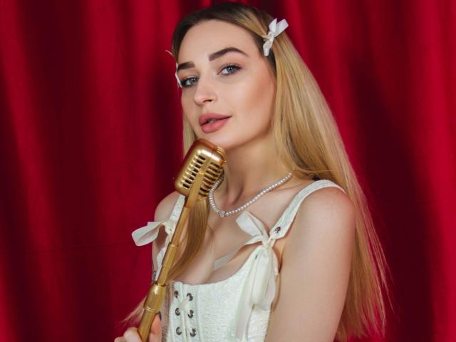 BarbaraRosy, una donna bionda con lunghi capelli, un fiocco bianco e una collana di perle, tiene un microfono dorato, indossando un corpetto bianco. con dettagli a forma di nastro, sfondo di tenda rosso, occhi azzurri, sguardo seducente, leggero sorriso.