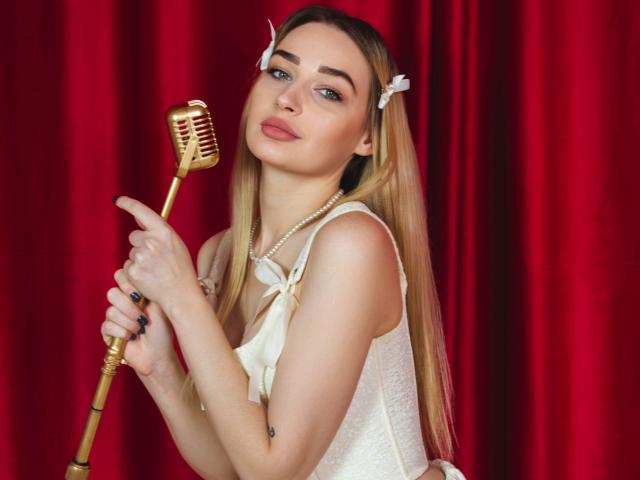 Barbara Rosy houdt een vintage gouden microfoon vast, ze draagt een witte kantjurk en een parelketting, met lang blond haar en een witte bloemclip. Rode gordijnen op de achtergrond. Sensuele, aantrekkelijke uitdrukking..