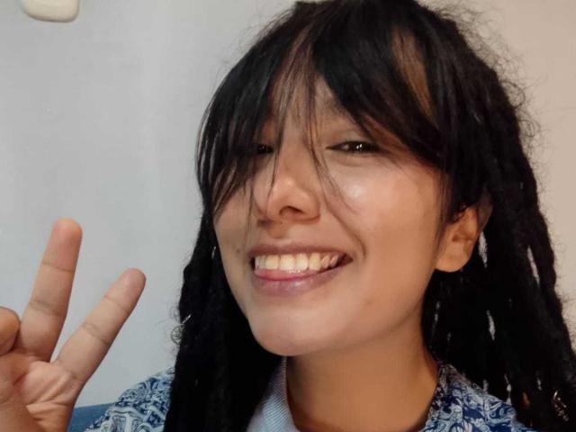 Gros plan de BeckyHuston, une jeune femme aux longs cheveux noirs, souriant avec les dents apparentes, faisant un signe de la paix. main droite.
sa peau est de couleur marron clair, et elle porte une chemise bleue à motifs..