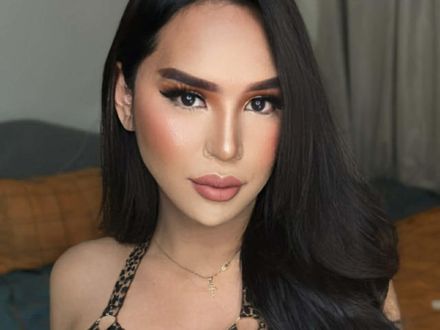 Close-up of SavanaStallionCock, a sexy woman with long black hair, heavy makeup, and a gold cross necklace. She has blue eyes, full lips, and a seductive gaze. Her top is leopard print.