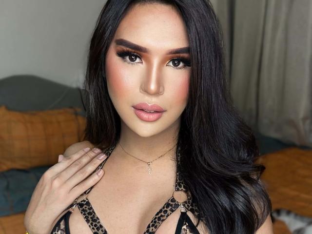 Close-up photo of SavanaStallionCock, a sexy woman with long black hair, heavy makeup, and full lips. She's wearing a leopard-print top, hand on chest, dark eyebrows, seductive gaze. Bright lighting.