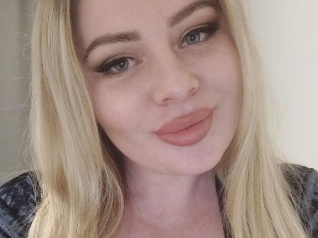 Gros plan de YourCoronet, une femme blonde à la peau claire, aux yeux bleus et aux lèvres roses et pulpeuses. Elle a des sourcils épais et porte du maquillage sombre pour les yeux. Son expression est légèrement séduisante..