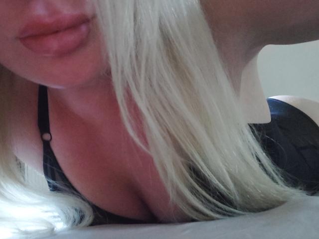 Close-up foto van het gezicht en de borst van YourCoronet, liggend op een bed, met blond haar dat gedeeltelijk hun zwarte beha bedekt. Grote decolleté. Lichtbron van onderen links, lippen lichtjes open..