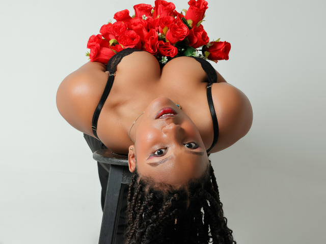 Upside-down photograph of TheaBarker, a dark-skinned woman with large breasts, wearing a black strapless top, holding red roses over her chest. Shiny skin, braided hair, intense gaze, seductive pose.