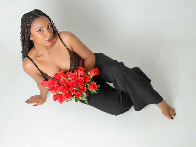 Thea Barker, uma mulher negra atraente com cabelo trançado, apoia-se no braço direito, vestindo uma blusa preta de renda e calças pretas. calças, com flores vermelhas vibrantes em um fundo branco. Muito atraente..