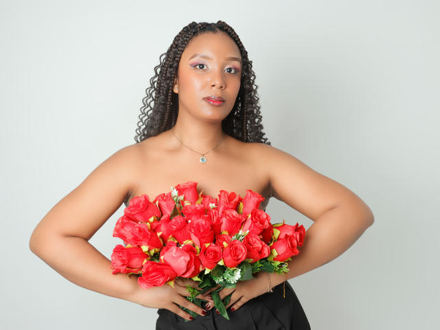 Fotografie einer schwarzen Frau mit lockigem Haar, die einen großen Strauß leuchtend roter Rosen vor einem schlichten weißen Hintergrund hält. Hintergrund. Sie trägt ein schwarzes Kleid ohne Träger und hat die Hände in die Hüften gestemmt. Ihr Ausdruck ist neutral, mit einem subtilen Hauch von Verführung..