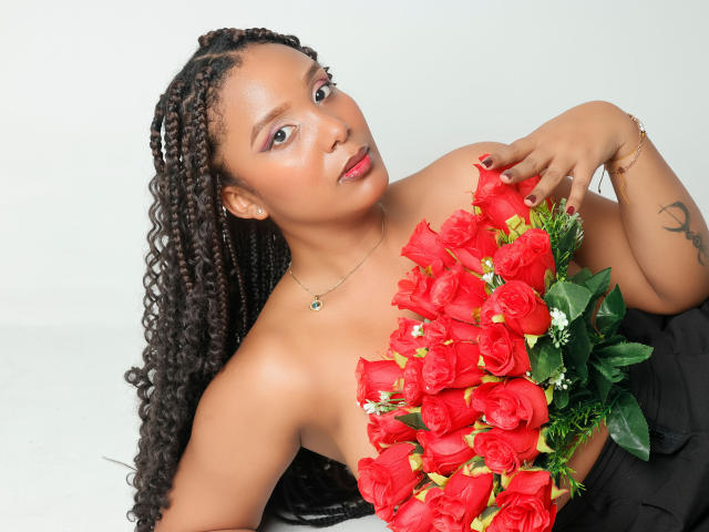 Photograph of TheaBarker, a curvy Black woman with long braids, holding a bouquet of red roses covering her chest. She gazes at the camera with a slightly open mouth. White background.