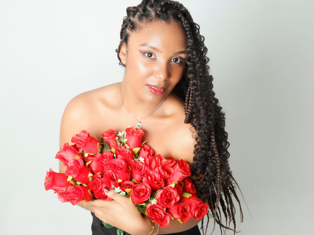 Photograph of TheaBarker, an attractive Black woman with long braids, holding a bouquet of bright red roses, covering her chest. She has a seductive expression, with intense blue eyes.