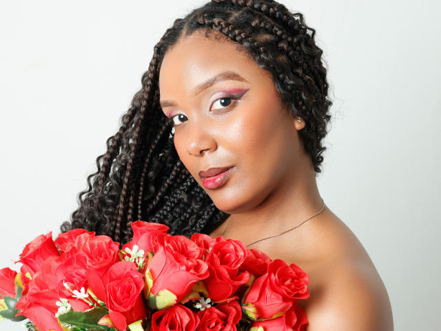 Photograph of a Black woman with long, curly hair holding a bouquet of bright red roses. She has a seductive gaze and a subtle smile. Her skin is shiny and smooth. Her breasts are partially visible. The roses are in the foreground.