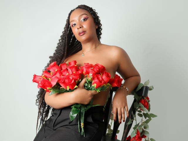 Fotografía de Thea Barker con piel oscura, cabello negro largo y rizado, sosteniendo rosas rojas brillantes que cubren su pecho. Ella lleva una falda negra y está apoyada en una silla, con más rosas. Increíblemente atractivo/a..