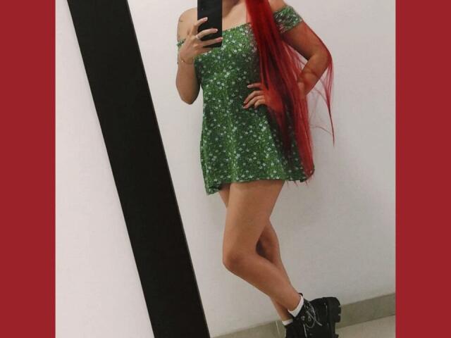 LaiaJonnes stands in front of a mirror, holding a phone, wearing a green sequined dress, black ankle boots, and long red hair. Her right hand rests on her hip, showing a confident pose.