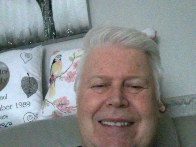 Foto em close-up de Stockings69, um homem mais velho com cabelo branco, sorrindo. O fundo inclui almofadas com estampas florais. A luz projeta sombras suaves, criando uma atmosfera relaxante e íntima. Expressão de felicidade extrema..