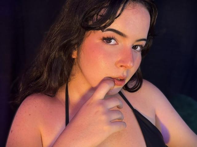 Close-up van AliinaFontaine, met een lichte huid en donker haar, die aan haar vinger knabbelt terwijl ze verleidelijk in de camera kijkt. Ze draagt een zwarte top met een halterhals, waardoor haar kleine borsten benadrukt worden. Warme, intense verlichting.