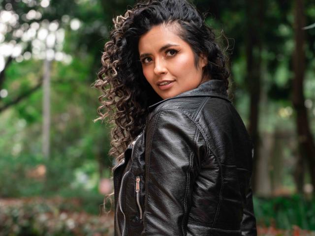 Fotografía de JedaZore, una mujer atractiva con cabello negro rizado, mirando por encima del hombro, vestida con cuero negro. chaqueta. Tiene una mirada seductora, con vegetación borrosa en el fondo..