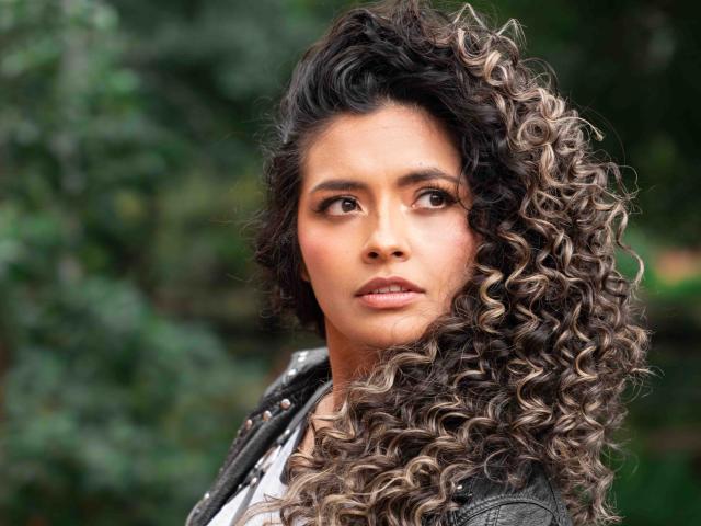 Fotografía de JedaZore, una mujer atractiva con cabello rizado, castaño y rubio, que mira directamente a la cámara con una expresión seria. expresión. Ella lleva una chaqueta negra sobre una camisa blanca..