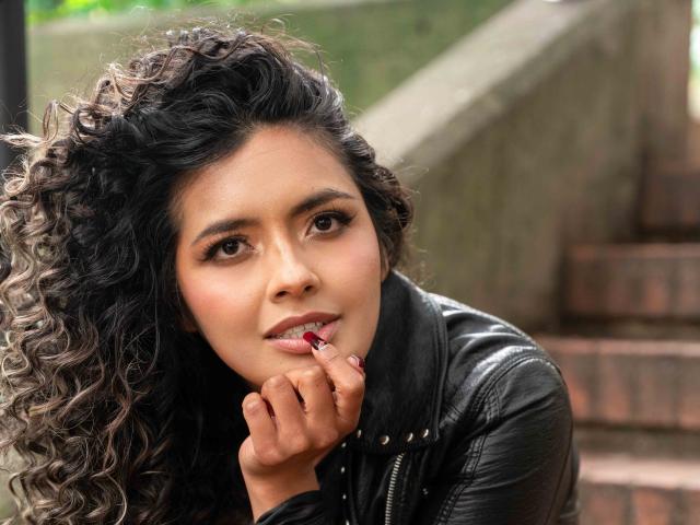 Photograph of JedaZore with curly black hair, dark skin, and brown eyes, biting her red-painted fingernail while wearing a black leather jacket. Blurred stairs in the background. Sexual tension evident.