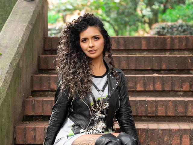 Photograph of JedaZore with long, curly brown hair, sitting on rusty metal stairs. Wearing a black jacket over a white and black patterned top. Slight, sexy smile. Background blurred greenery.