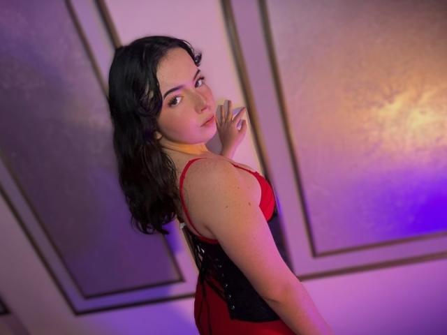Photograph of AliinaFontaine, standing in a dimly lit room with purple and blue lighting. She has wavy black hair, wearing a red and black dress, looking over her shoulder with a sultry expression.