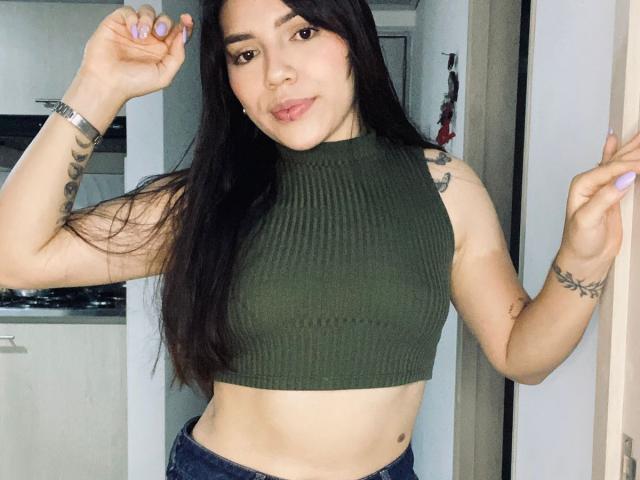 Photograph of SophiaMarx with long black hair, wearing a green ribbed crop top, exposing her midriff, posing with hands up, showing tattoos on arms, intense gaze, slightly parted lips.