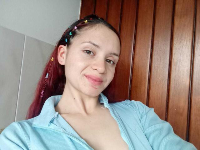 Lesley Gray, con el pelo largo y rojo, vestida con una camisa azul claro, apoyada contra un panel de madera. Está sonriendo ligeramente, y la luz natural resalta su piel clara. Pinzas para el pelo visibles. Estilo casual y sexy..