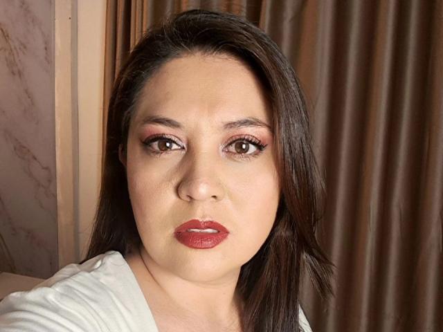 Close-up van PaulaAnd, een Latina vrouw met donkerbruin haar, rode lippenstift en bruine ogen. Ze draagt een witte blouse en kijkt recht in de camera met een enigszins uitdagende blik..