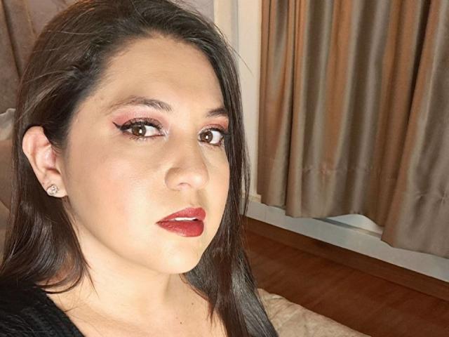 Close-up photo of PaulaAnd, a sexy woman with long black hair, red lipstick, and pink eye shadow, looking directly at the camera with a slight smile. Brown curtains in the background.