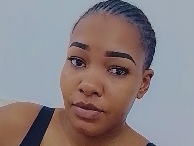 Close-up selfie van SexyApplexx, een aantrekkelijke zwarte vrouw met gevlochten haar, die een zwarte tanktop draagt. Ze heeft volle lippen, gebogen wenkbrauwen en een neutrale uitdrukking, met een lichtblauwe achtergrond..