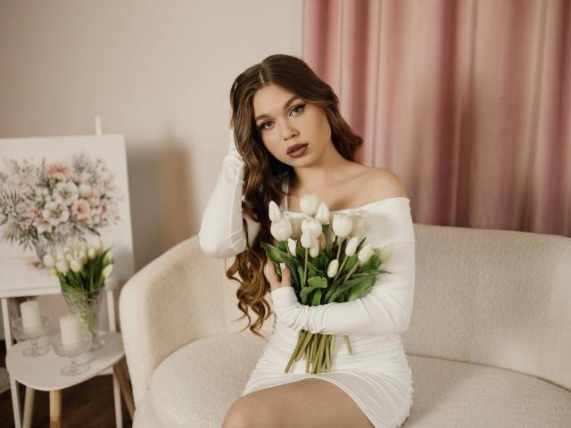 LaraPika, avec de longs cheveux bruns ondulés, assise sur un canapé blanc, portant une robe blanche à une épaule, tenant un bouquet de des tulipes blanches, des rideaux roses en arrière-plan, expression sexy et aguicheuse..