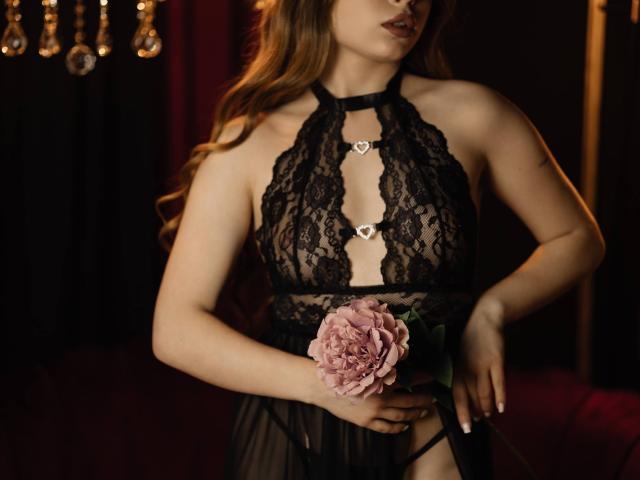 LaraPika with long brown hair wears a black lace halter top, exposing cleavage and heart-shaped cutouts. Holds a pink flower, posing seductively, dim lighting, shadows accentuating her curves.