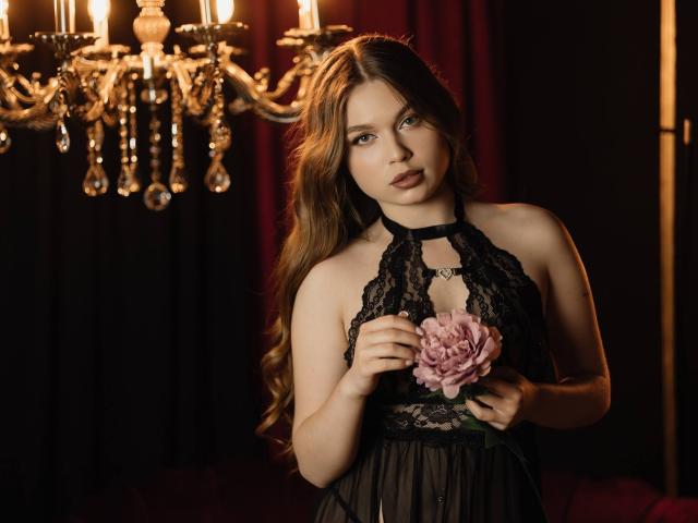 LaraPika aparece en el centro del encuadre, sosteniendo una flor rosa y vestida con un vestido negro de encaje con escote halter. Ella tiene el pelo largo y castaño y una expresión seductora. Un candelabro brilla detrás de ella..