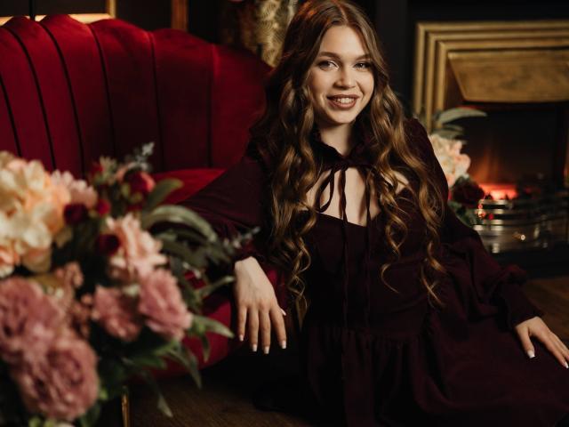 LaraPika, com longos cabelos castanhos ondulados, sorrindo, vestindo um vestido preto com um laço, sentada em um sofá de veludo vermelho, rodeado por flores rosa e brancas, e uma lareira acesa..