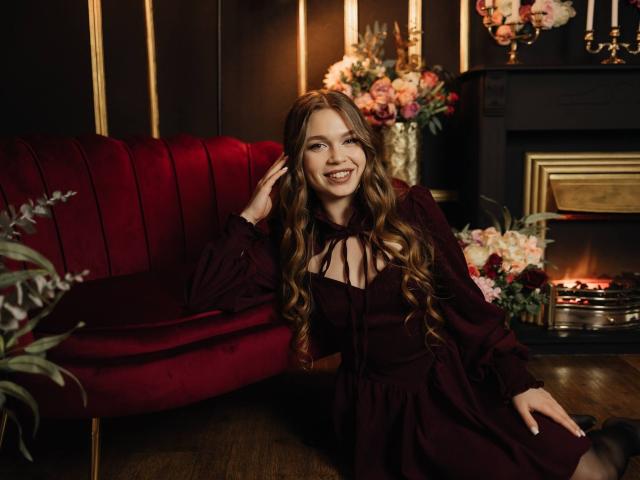 LaraPika, una mujer sonriente con el pelo largo y castaño, está sentada en un sofá de terciopelo rojo, vestida con un vestido negro con un detalle de lazo. El fondo incluye una chimenea encendida, flores y paneles oscuros..