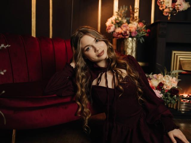 LaraPika leans back against a red velvet couch, long wavy brown hair, black dress with sheer neckline, seductive gaze, dimly lit room, fireplace with pink flowers, soft light, sexy vibe.