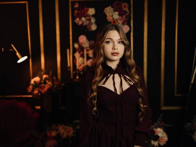 LaraPika steht in der Mitte, trägt ein schwarzes Kleid mit Halstuch, hat langes, gewelltes braunes Haar und einen intensiven Gesichtsausdruck. Dunkler, floraler Hintergrund mit weicher, oranger Beleuchtung. Verdammt sexy..