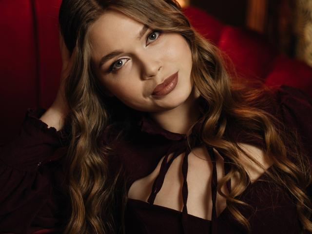 Close-up photo of LaraPika with long, wavy brown hair, wearing a black strappy top, looking directly at the camera with a slight smile, red lighting in the background, sexy.