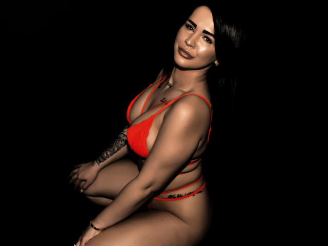 SavageSya, com longos cabelos pretos, vestindo um biquíni vermelho, agachada com uma tatuagem no braço esquerdo, olhando para trás. Expressão sensual, fundo preto, destacando seu corpo curvilíneo e sexy..