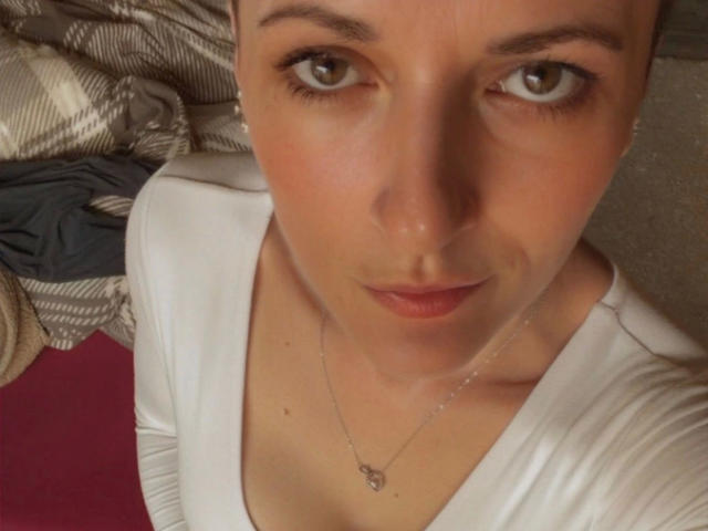 Primer plano de NastyMommy, mostrando su belleza, con énfasis en su rostro y parte superior del cuerpo. Ella tiene el pelo corto y castaño, ojos marrones, y lleva una camiseta blanca con cuello en V y un collar delgado. Iluminación suave. Increíblemente sexy..