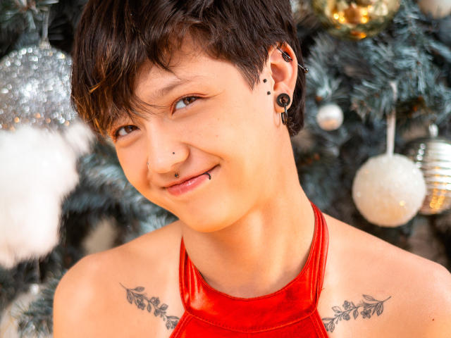 Close-up photo of LissyLuxe, a young person with short black hair, smiling, wearing a red halter top, black ear gauges, and shoulder tattoos. Blurred Christmas tree with white ornaments in background.