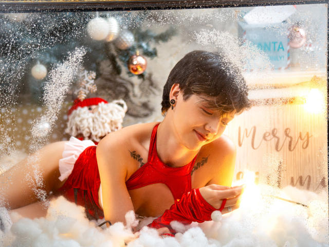 LissyLuxe with short black hair, red lingerie, and white fur, lying in a snow-like substance, gazing downwards with a seductive expression, glowing light behind her, blurred Christmas hat in background.